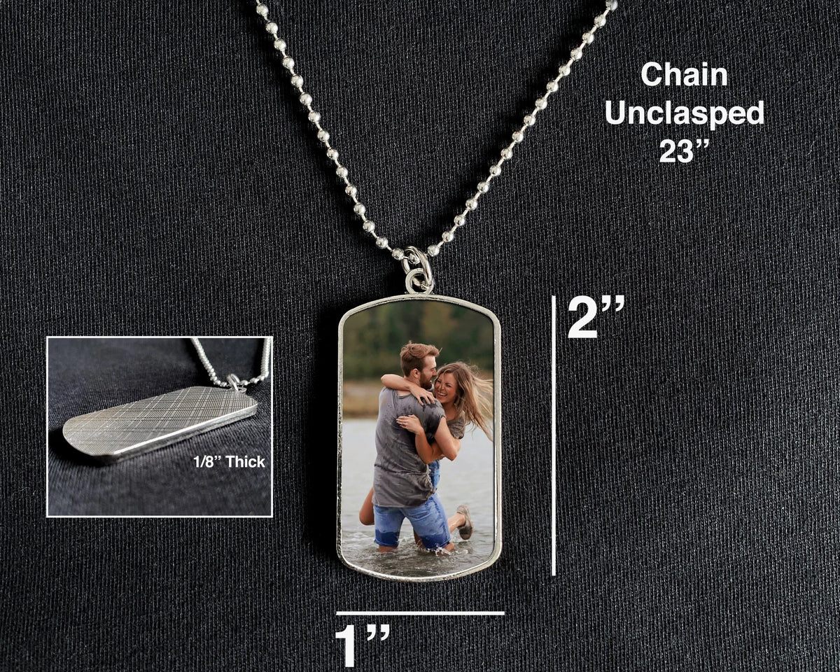 A silver dog tag necklace with a custom photo insert, shown on a black textured background with dimension labels indicating 2 inches tall by 1 inch wide, plus a small inset showing the unclaspable chain and 1/8 inch thickness.