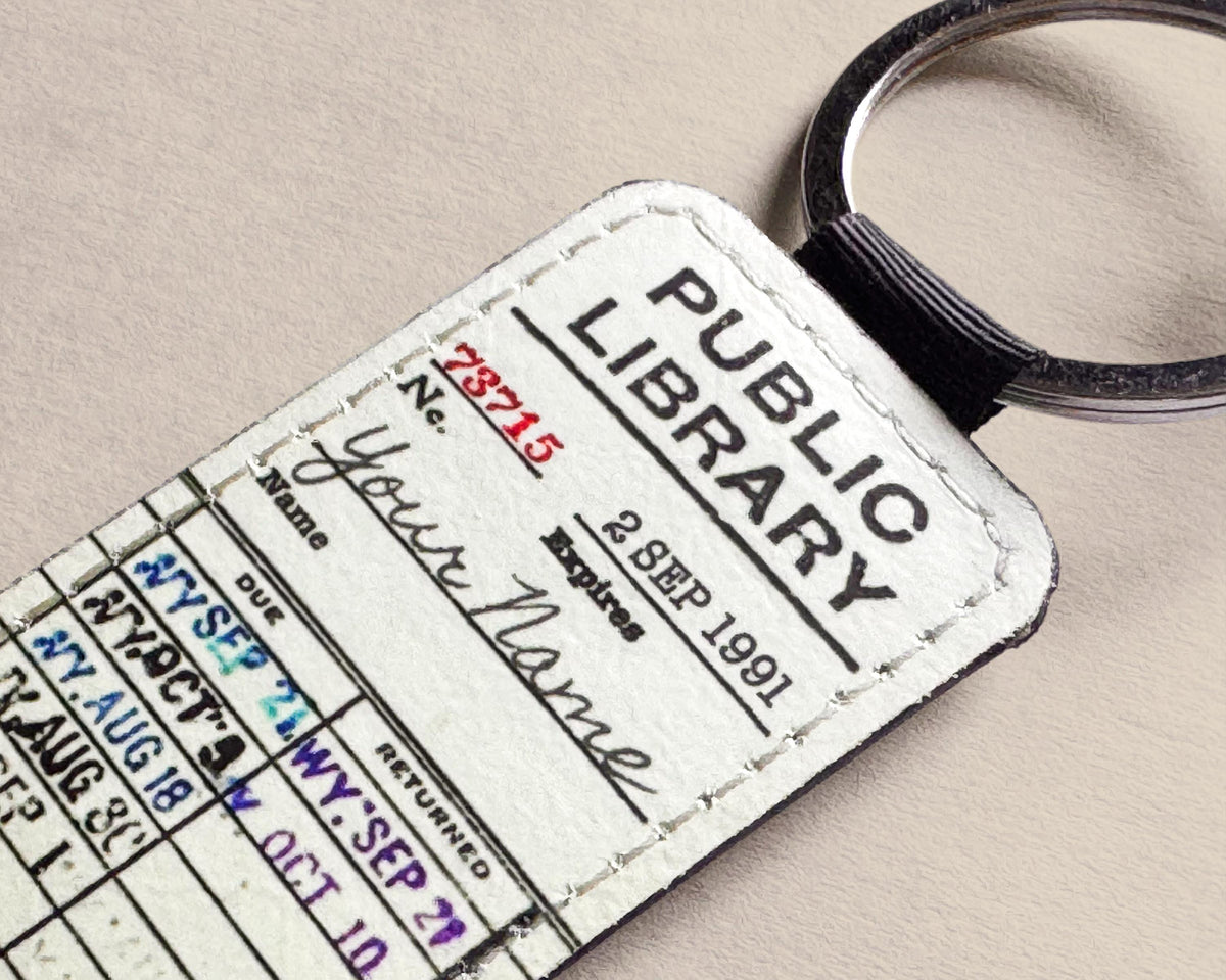 A novelty keychain shaped like a vintage library card labeled 'PUBLIC LIBRARY,' with handwritten-style details and a black elastic loop attached, shown on a light beige textured background.