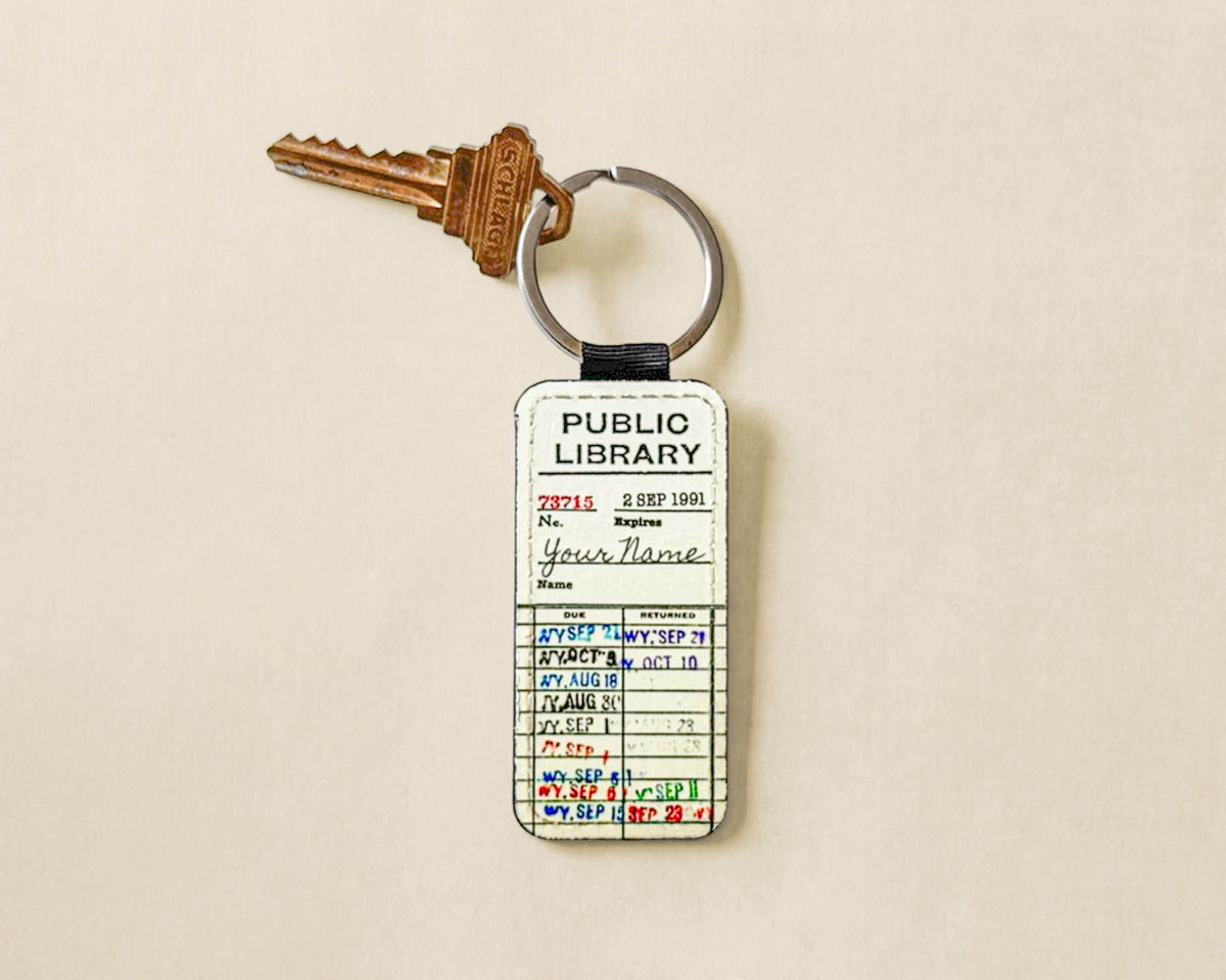 A novelty keychain featuring a vintage library badge card with handwritten details, attached to a split keyring with a brass key, set against a light beige background.