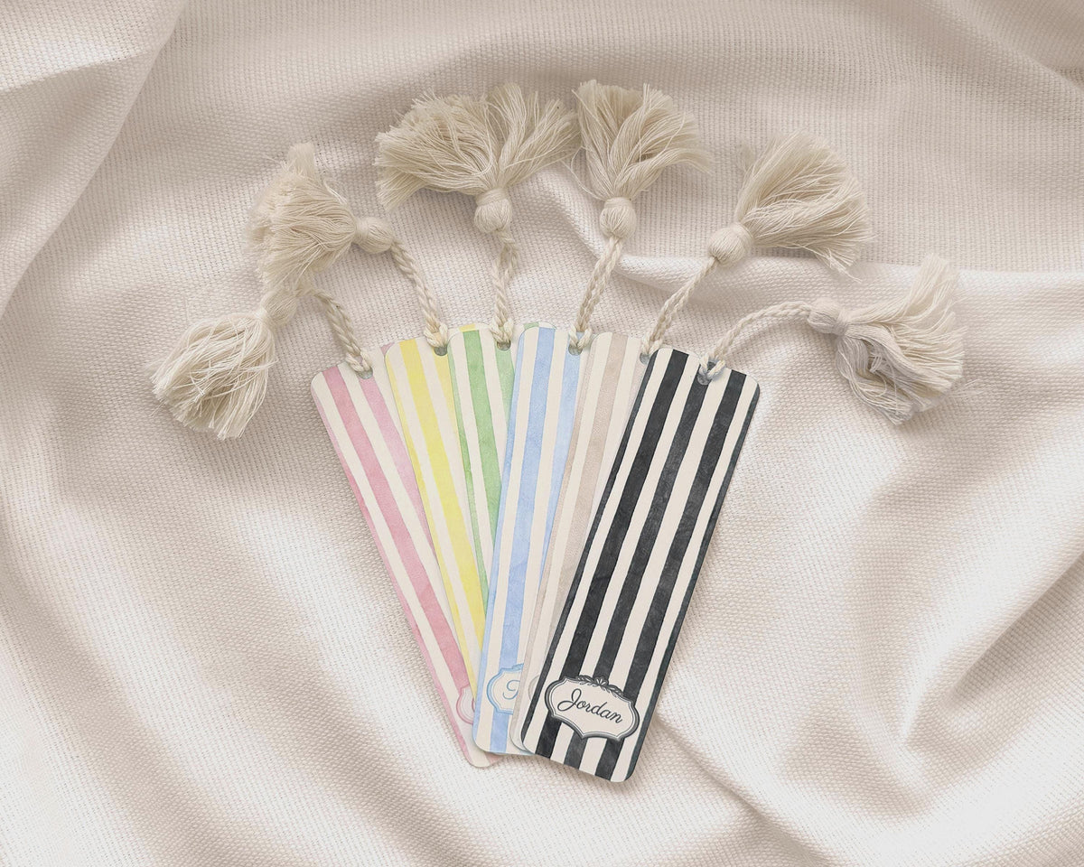 Four decorative metal bookmarks with cream tassels, arranged on a draped cream fabric background. The bookmarks feature vertical stripes in pink, yellow, green, blue, black, and white, with one bookmark personalized with the name 'Jordan'.