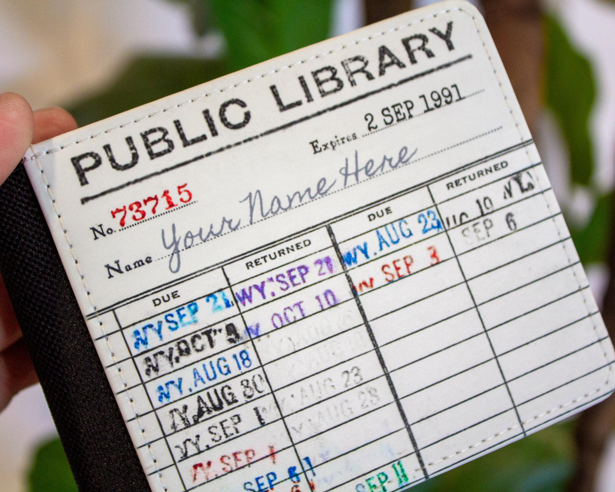 A close-up of a novelty passport-style holder or wallet being held by a hand. The front is printed with a vintage library form reading 'PUBLIC LIBRARY' and 'Your Name Here,' with sample stamped dates in red and blue ink and a black fabric band on the left.