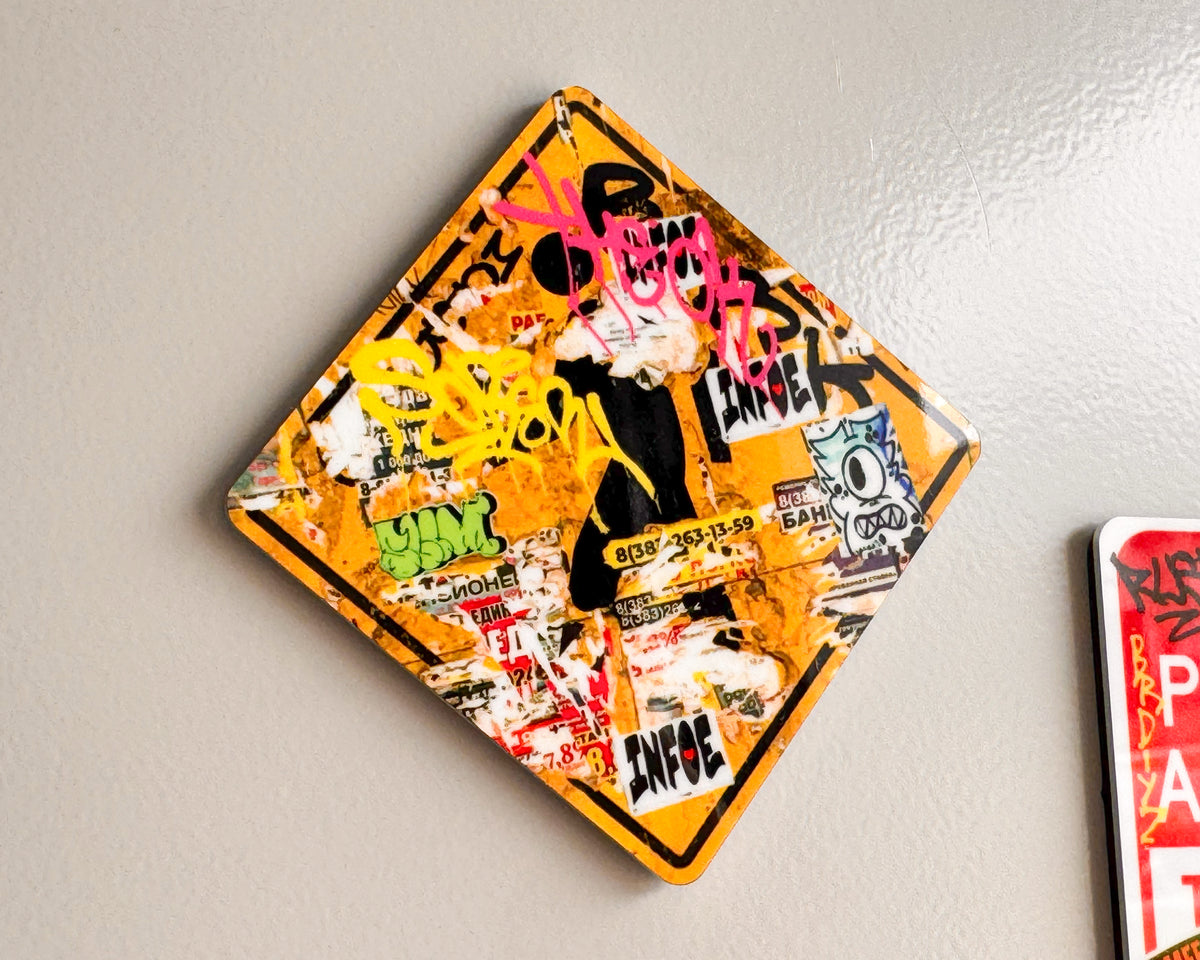 A diamond-shaped graffiti fridge magnet featuring a distressed yellow warning-style border with layered graffiti, stickers, and bold black and pink lettering, mounted on a light gray wall.