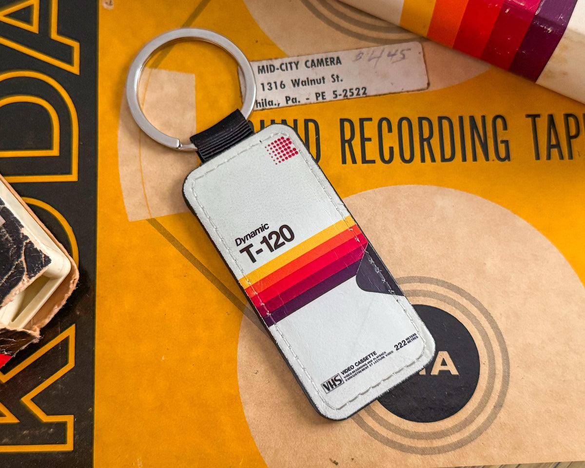 A vintage-style keychain with a silver split ring and a rectangular tag designed to look like a Dynamic T-120 cassette tape, featuring a retro striped color block pattern in red, orange, yellow, and purple on a light gray background, displayed on a vintage record sleeve.