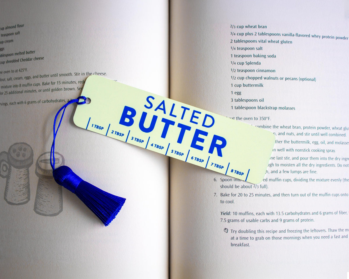 A rectangular light yellow recipe bookmark with blue text reading 'SALTED BUTTER' and a blue string, laid on top of an open cookbook page.