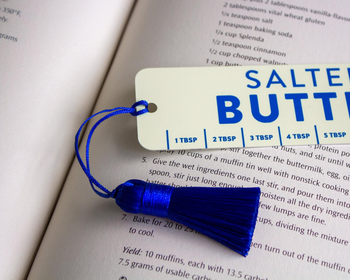 A blue tassel salted butter bookmark with a yellow color 'SALTED BUTTER' and tablespoon markings, placed over an open recipe book.