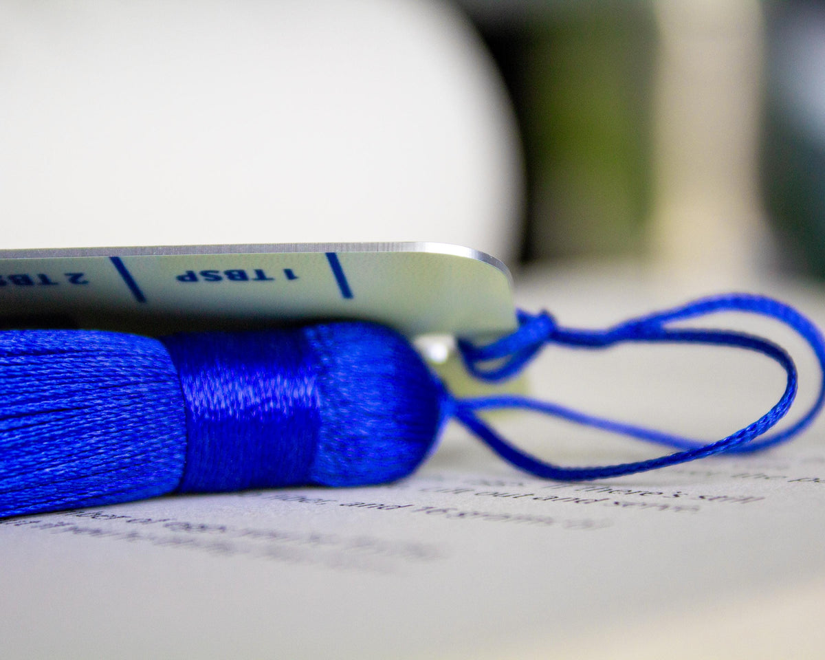 A close-up of a bright blue silk bookmark with a tassel and loop, resting on an aluminum salted butter bookmark