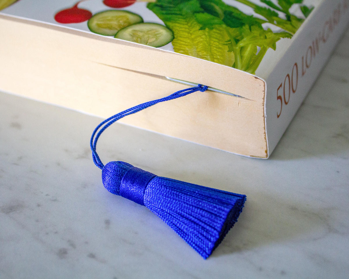 A bright blue fabric bookmark with a matching tassel is threaded through the edge of a book on a light marble surface.