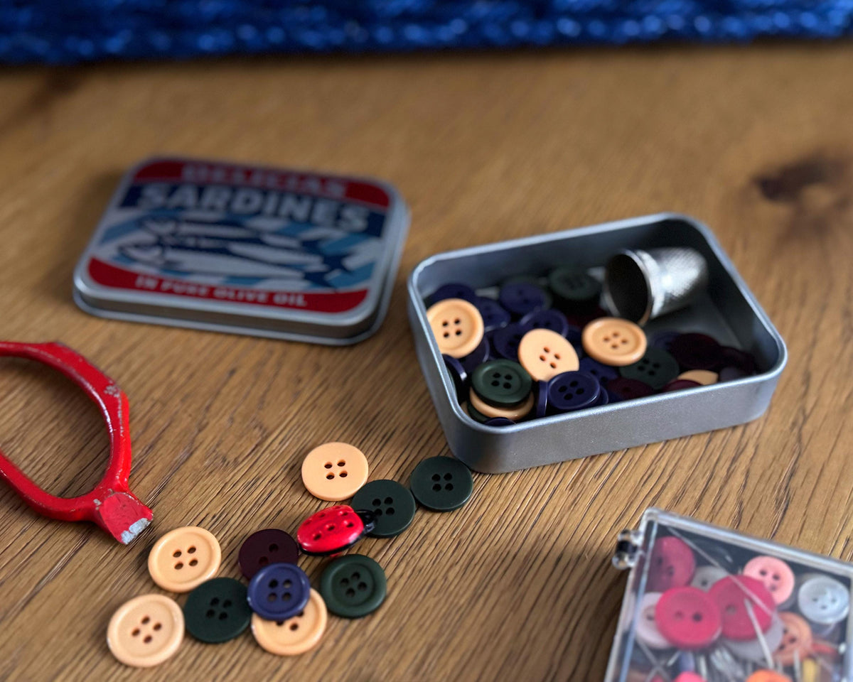 A sardines tin of sewing buttons and a small metal thimble on a wooden tabletop, with loose multicolored buttons scattered nearby and a red-handled pin or needle holder on the left.