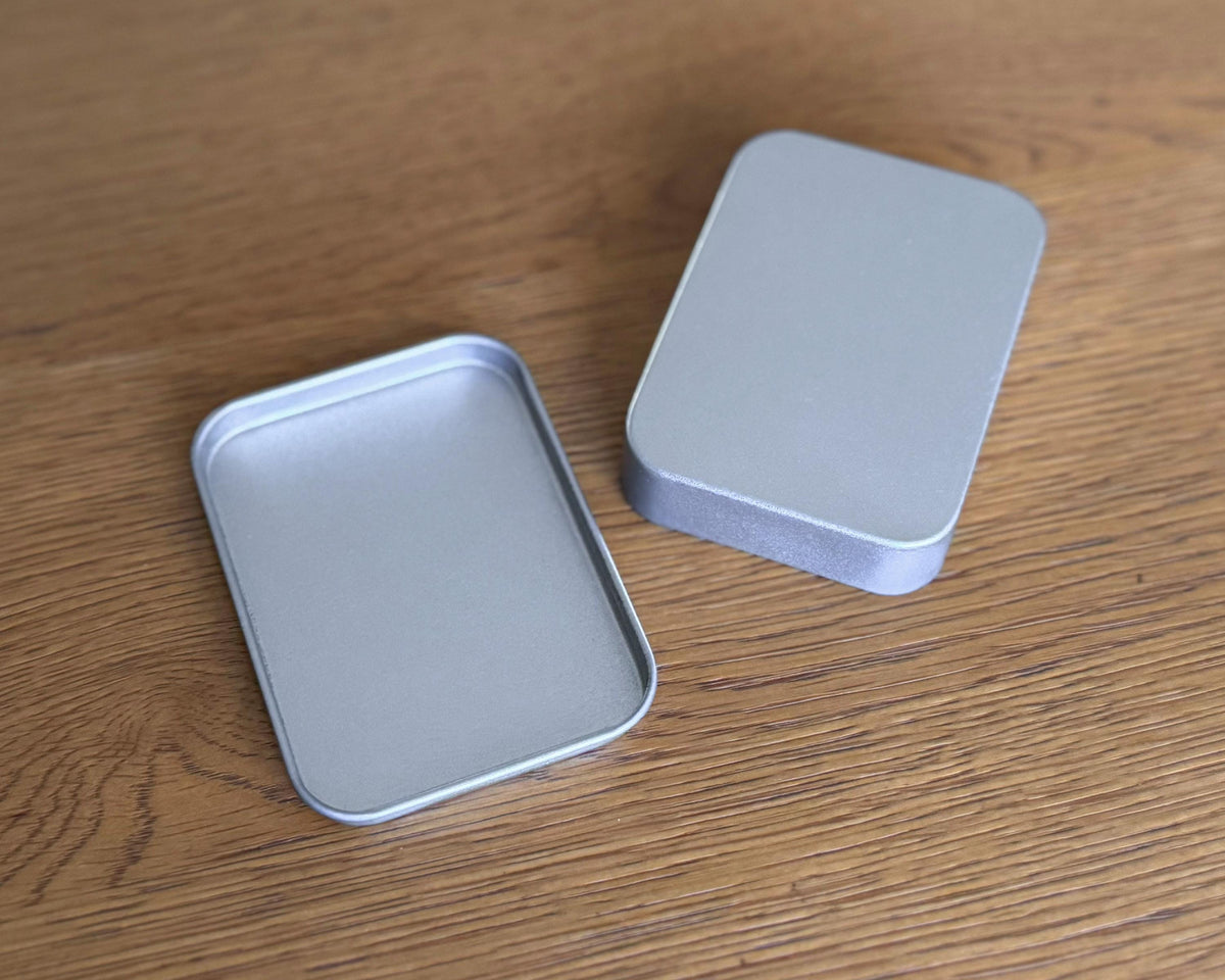 Two empty rectangular silver metal tins with rounded corners, one open and one closed, placed on a wooden surface.
