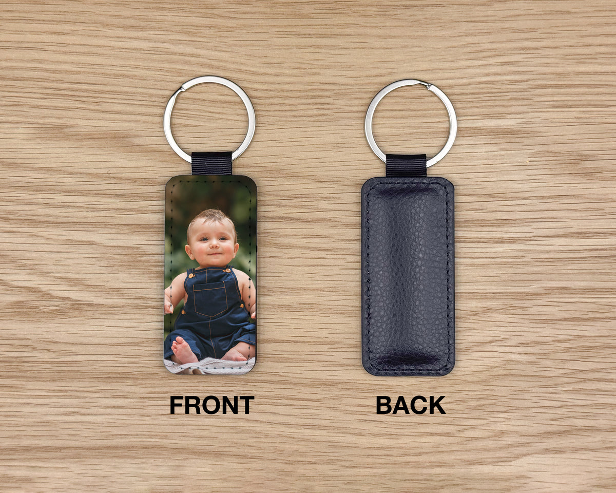 Two views of a custom rectangular keychain on a light wood background. The left 'FRONT' view shows a photo of a baby in a blue outfit, and the right 'BACK' view shows a dark textured leather back.
