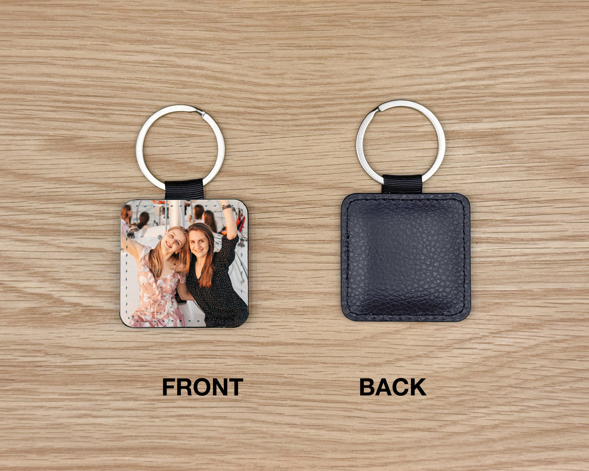 Two square leather keychains on a light wood background, labeled FRONT and BACK. The left keychain shows a printed photo insert, and the right keychain shows the dark navy pebbled leather back with a silver key ring.