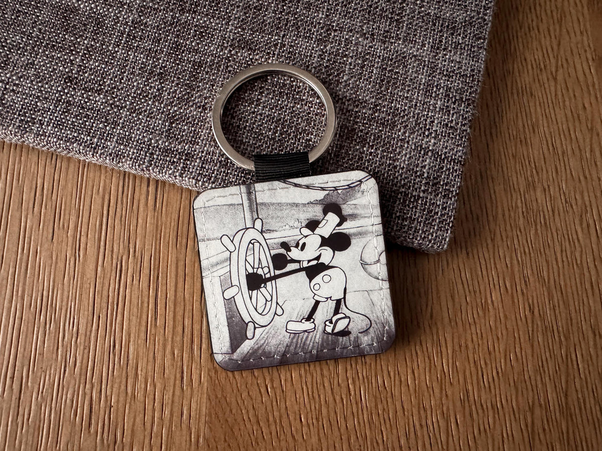 A square vegan leather  keychain featuring a black and white illustration of a mouse at the ship's wheel, resting on a textured gray fabric over a wooden surface.