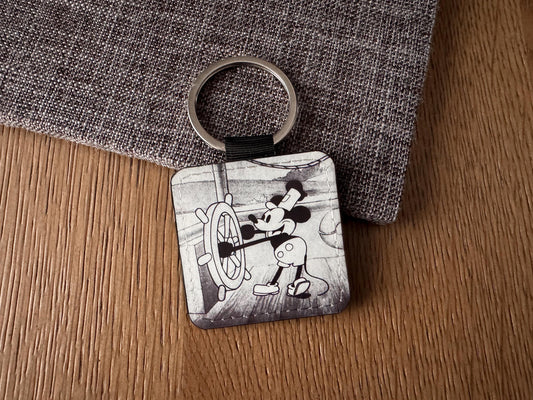 A square vegan leather  keychain featuring a black and white illustration of a mouse at the ship's wheel, resting on a textured gray fabric over a wooden surface.