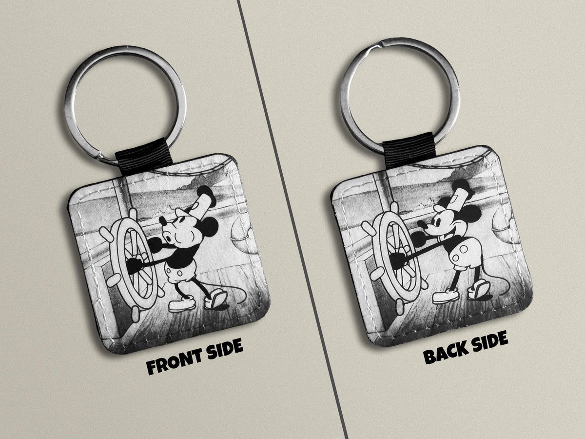 A square keychain shown from both front and back sides on a light beige background. The keychain has a silver metal split ring and a black strap, with a black-and-white illustrated print of Steamboat Willie  at a ship's wheel on the face.