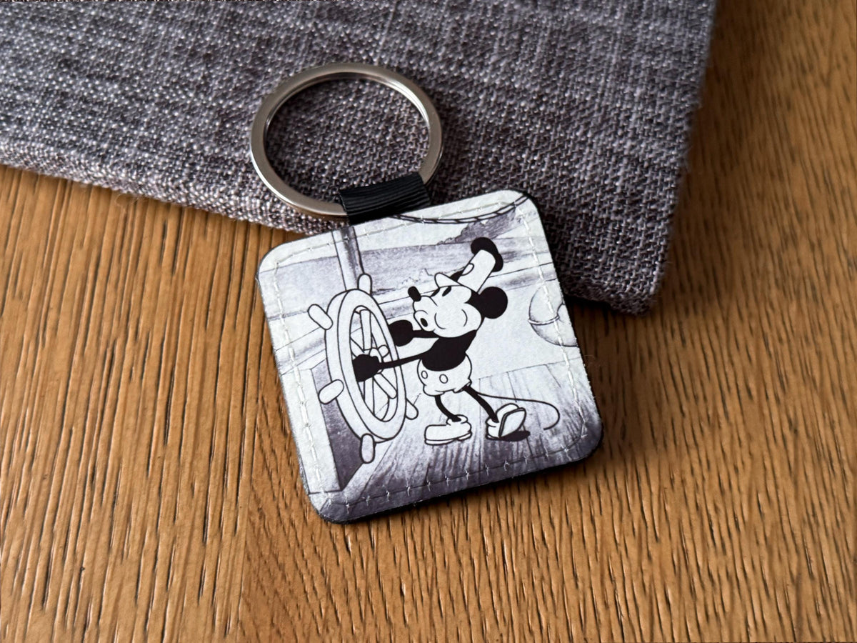 A square keychain featuring a black and white vintage steamboat  willie  illustration of a Mouse at the ship's wheel, resting on a wooden surface next to a gray textured fabric.