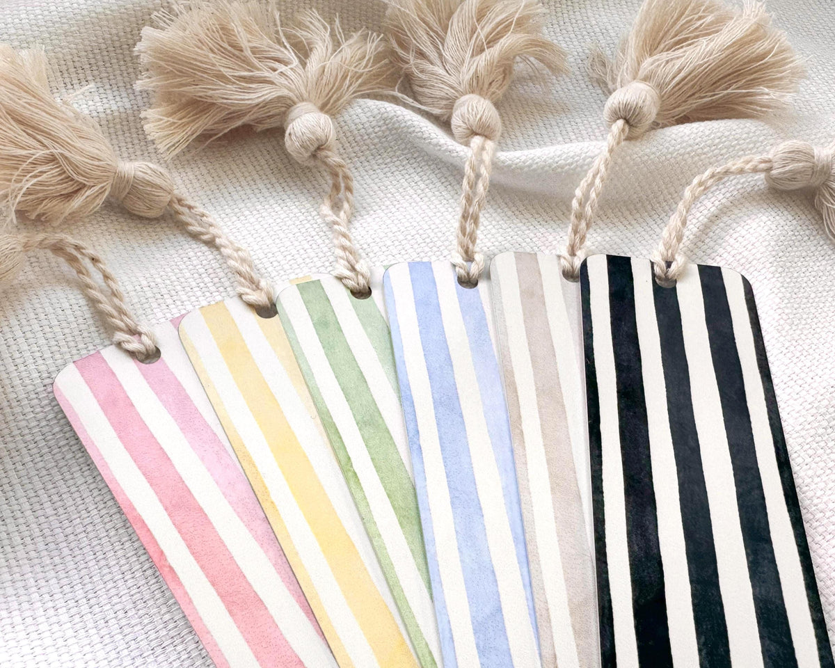 Six striped aluminum bookmarks with cream tassels, arranged in a fan shape on a textured white fabric background. The bookmarks feature vertical stripes in pink, yellow, mint green, light blue, beige, and black.