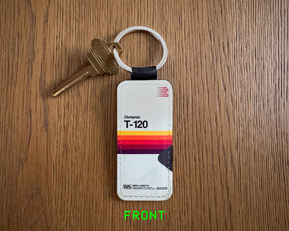 A vintage-style keychain with a white plastic tag featuring the words 'Dynamic T-120' and a retro stripe pattern, attached to a black strap and silver key ring, placed on a wooden surface with a brass key beside it.
