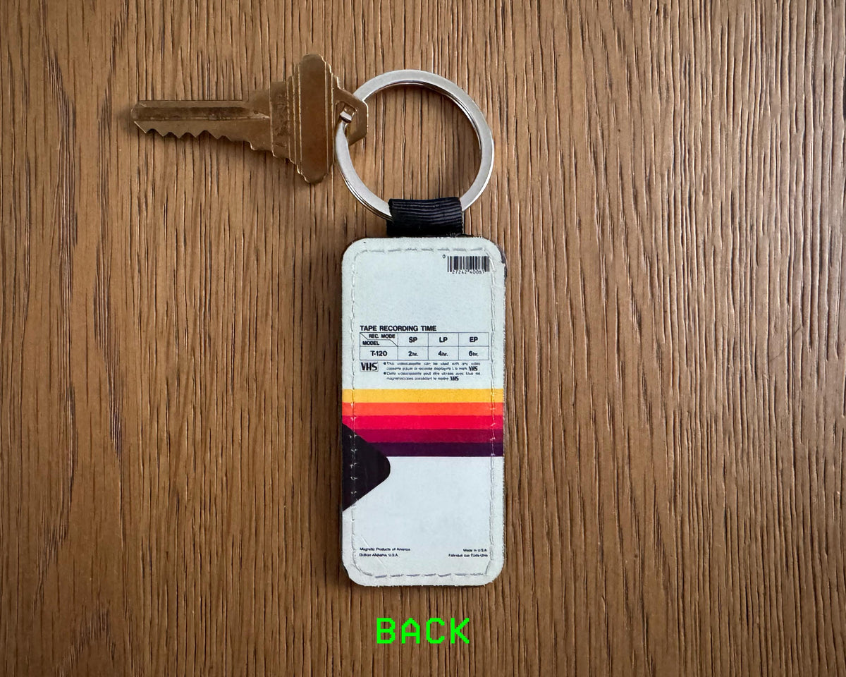 A vintage-style leather luggage tag keychain with a white front, a colorful retro stripe pattern in orange, yellow, and red, and a black card holder pocket, shown on a wooden table next to a key. The word 'BACK' is written in green at the bottom.