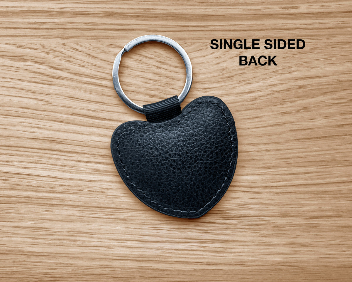 A black pebbled leather heart-shaped keychain with a silver metal split ring, shown on a light wood grain background with the text 'SINGLE SIDED BACK' in the upper right.