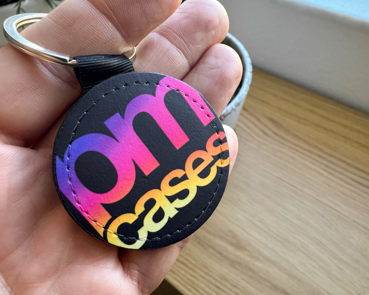 A close-up of a hand holding a round black keychain with a colorful gradient 'om cases' text design and a gold split ring.