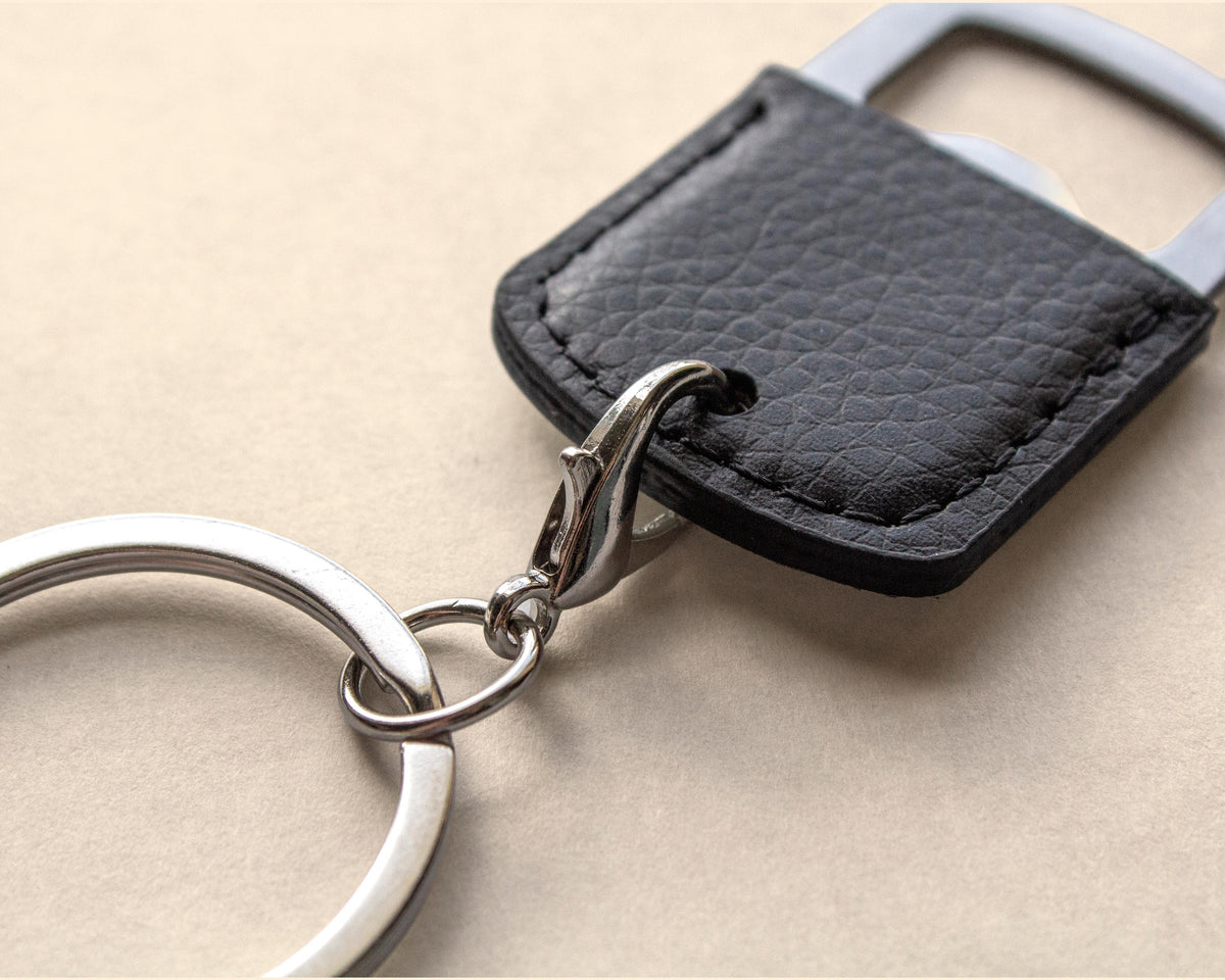 A close-up of a black textured leather keychain tag with a silver metal split ring and bottle opener attachment, resting on a neutral beige background.