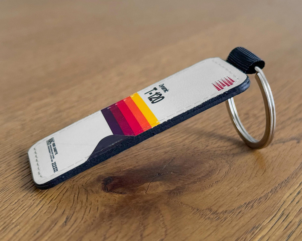 A close-up of a leather keychain tag styled like a credit card, featuring a white body, a colorful stripe graphic in red, orange, yellow, and purple, and a silver metal key ring, resting on a wooden surface.