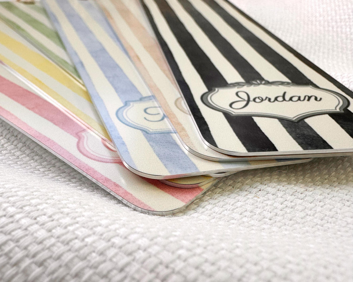 A close-up of several decorative aluminum bookmarks fanned out on a white textured fabric. The top tag has black and white stripes with a white label reading 'Jordan', while the others show pastel stripes in yellow, green, blue, and pink.