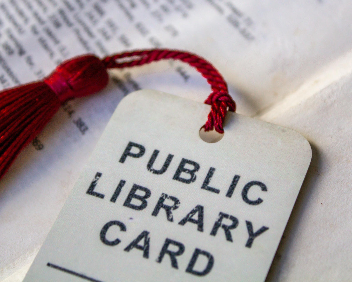 A close-up of a white aluminum public library card tag with a red tassel string, resting on an open book with printed pages visible in the background.