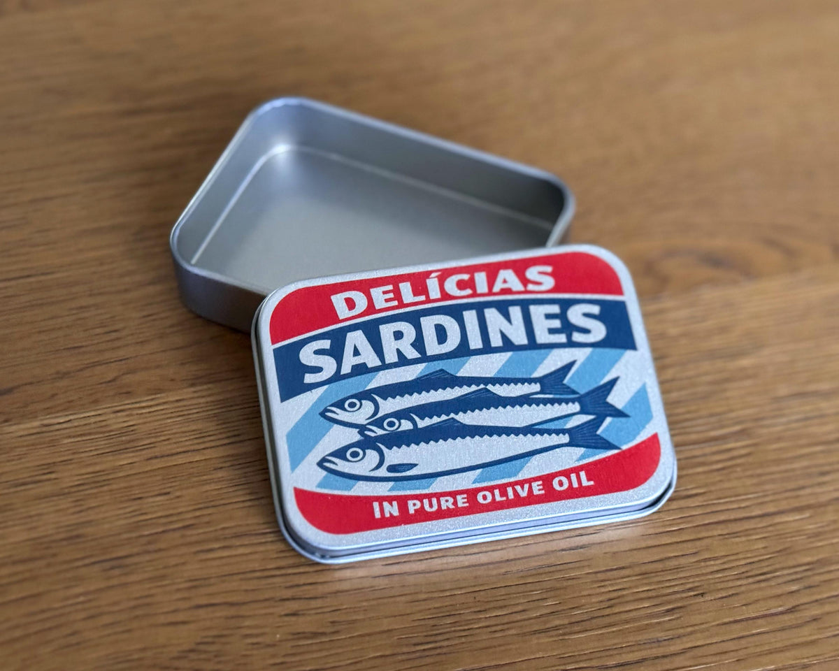 A square metal tin of Delicias Sardines in pure olive oil, shown open with its lid leaning against it on a wooden tabletop. The lid features a red, white, and blue label with illustrations of sardines.